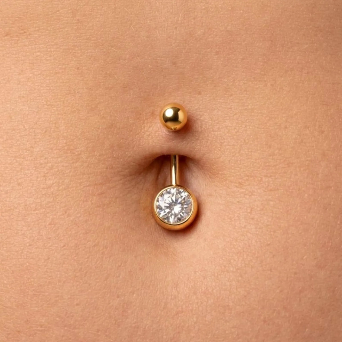 A close-up of a gold navel piercing stud with a clear round gemstone, shown on a person's skin.