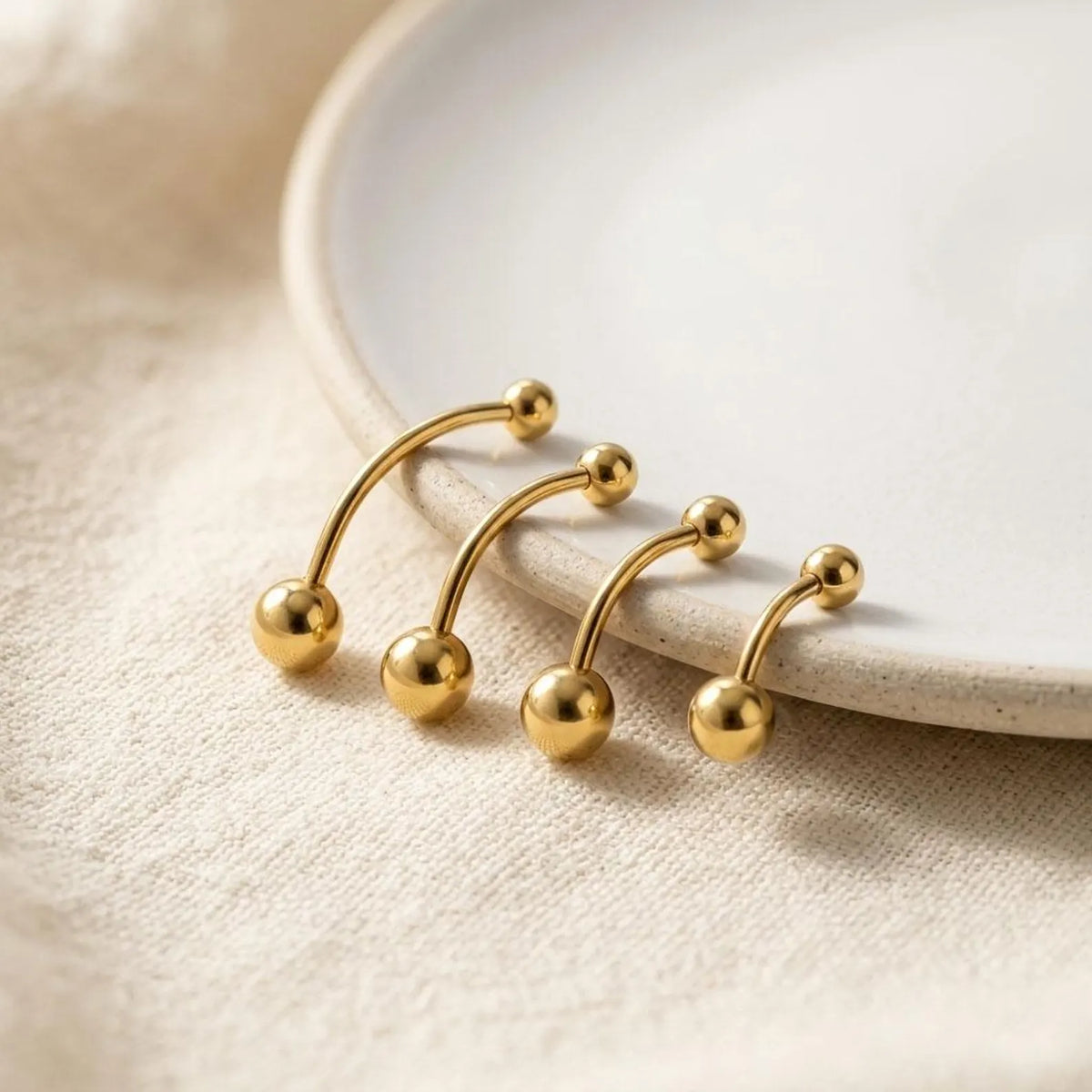 Four gold-plated curved barbell belly button piercings with spherical ball studs, arranged on a textured beige fabric next to a white ceramic plate.