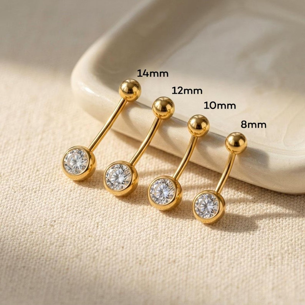 Four gold-toned barbell stud piercings with clear gemstones are arranged on a beige fabric surface beside a white dish, with size labels 14mm, 12mm, 10mm, and 8mm overlaid.