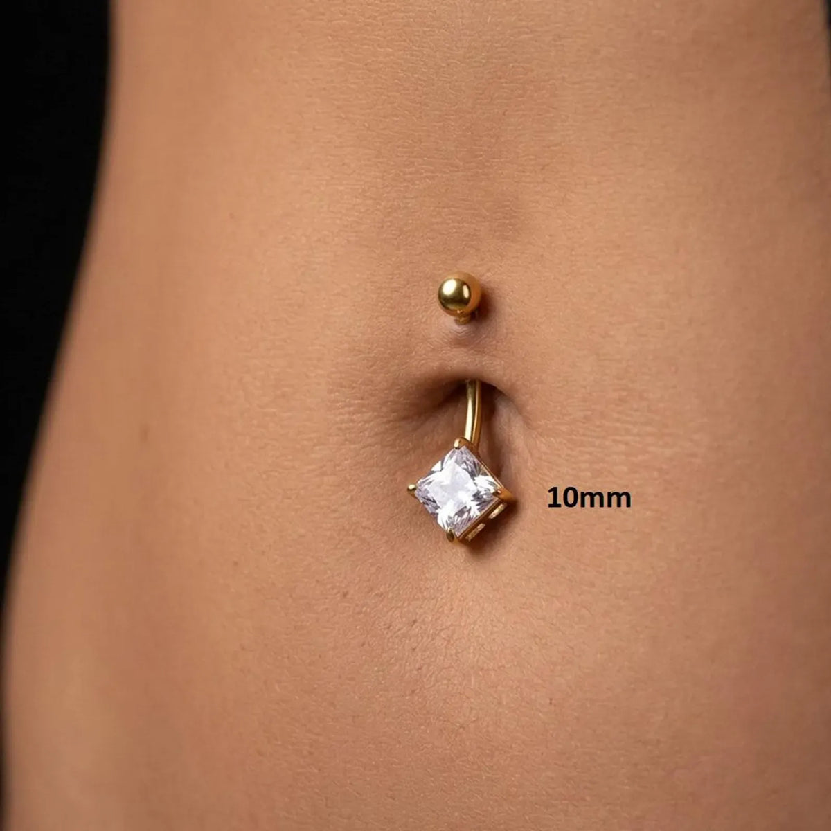 A close-up of a belly button piercing with a gold barbell and a clear square gemstone, shown on a person's abdomen with a '10mm' size marker.