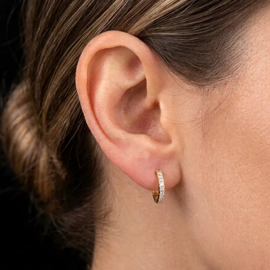 A close-up of a person's ear wearing a small gold huggie hoop earring set with clear stones, against a dark background.