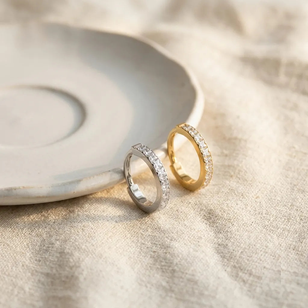 Two slim eternity rings, one in silver and one in yellow gold, both set with small clear stones, resting on a textured beige fabric next to a ceramic dish.