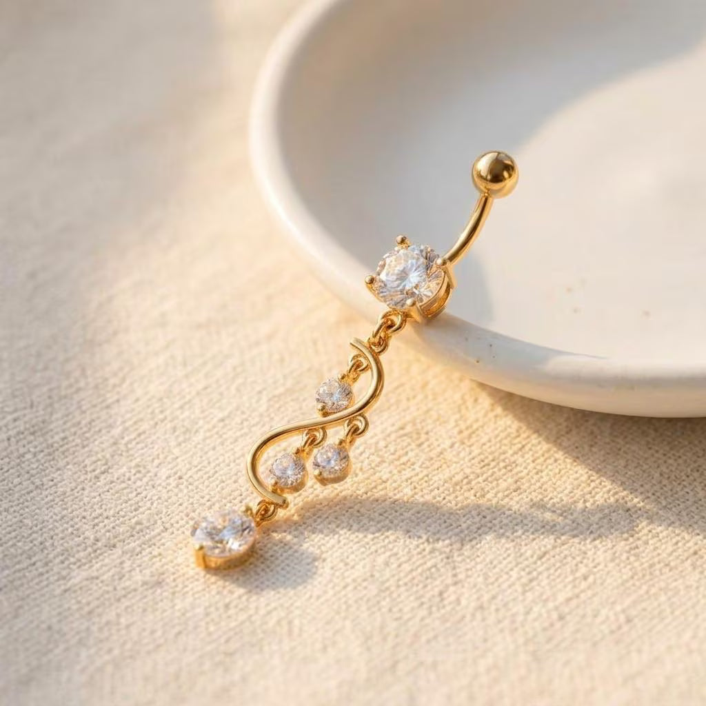 A gold-plated curved navel piercing barbell with multiple clear gemstones, resting on a textured beige fabric next to a white ceramic dish.
