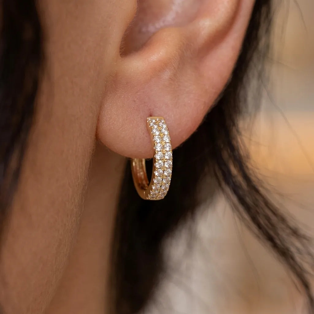 A close-up of a gold huggie hoop earring paved with small clear stones, worn on a person's ear.