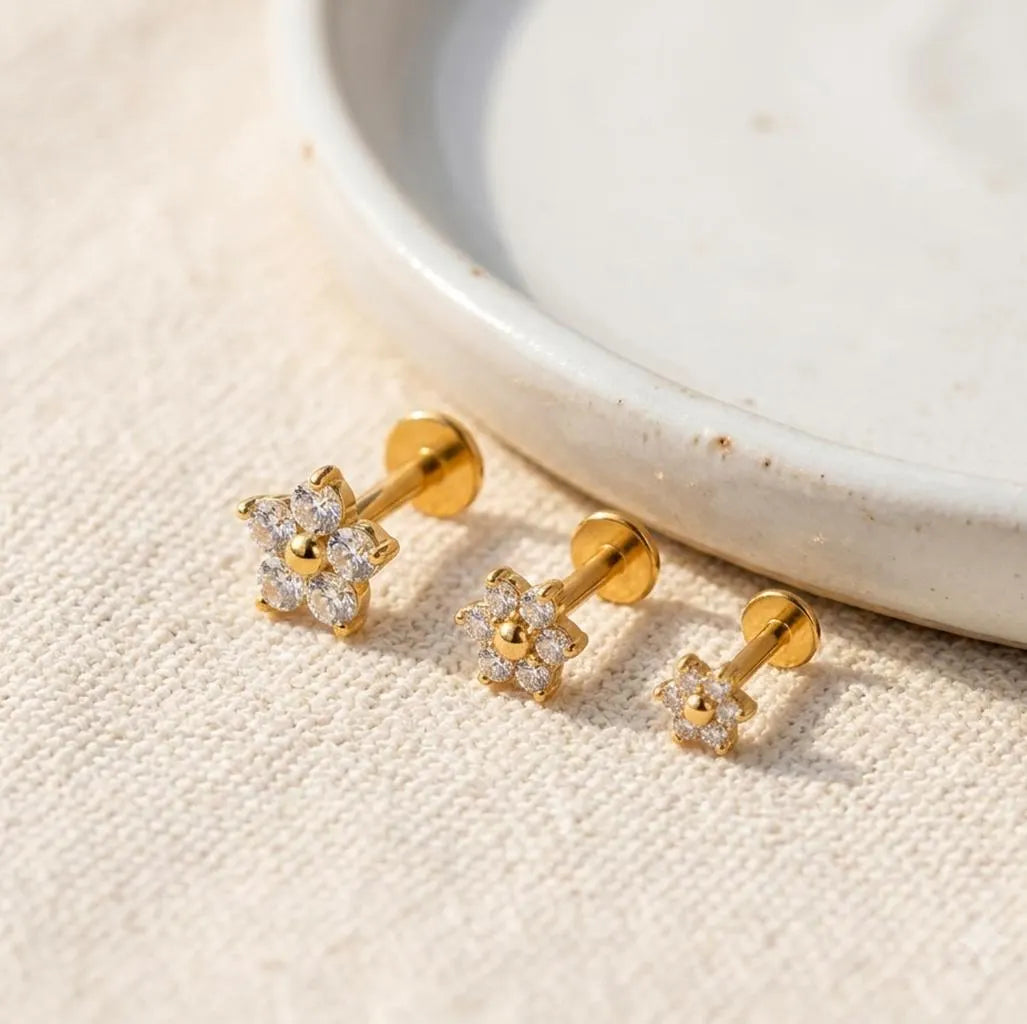 Three gold flower-shaped stud earrings with clear stones, arranged on a textured beige fabric next to a white ceramic dish.