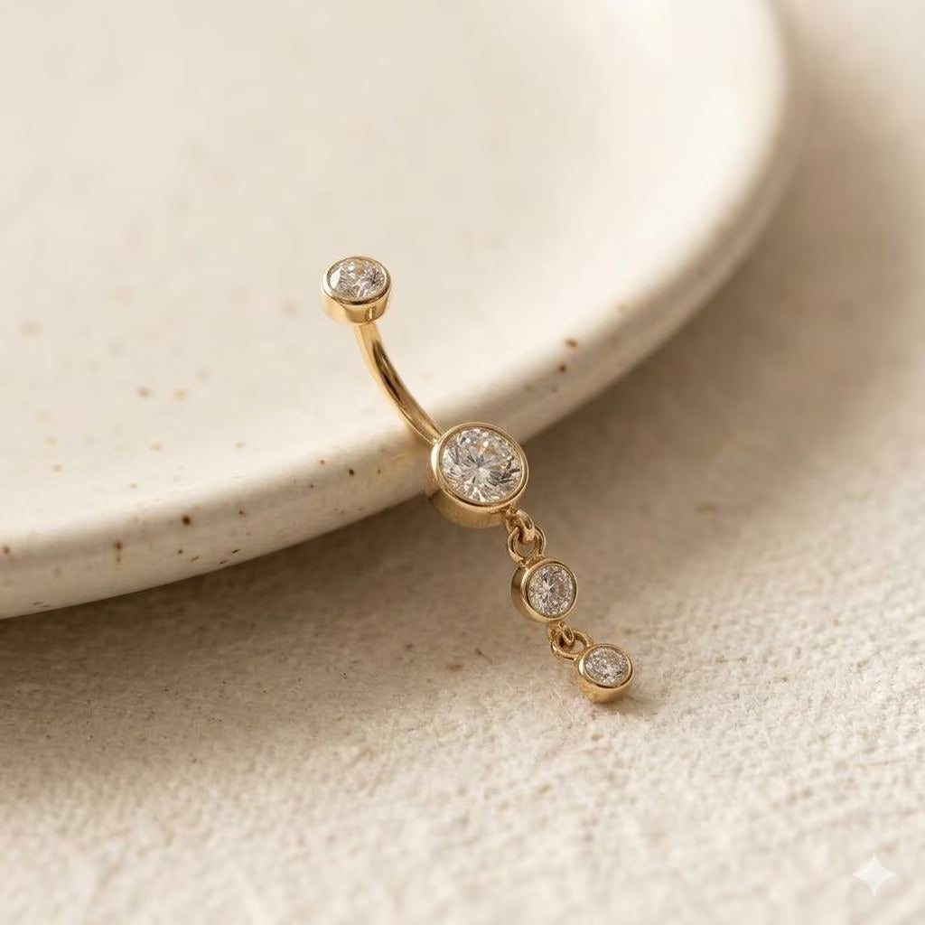 A gold curved barbell belly button ring with three dangling clear round gemstones, resting on a textured beige surface next to a ceramic dish.