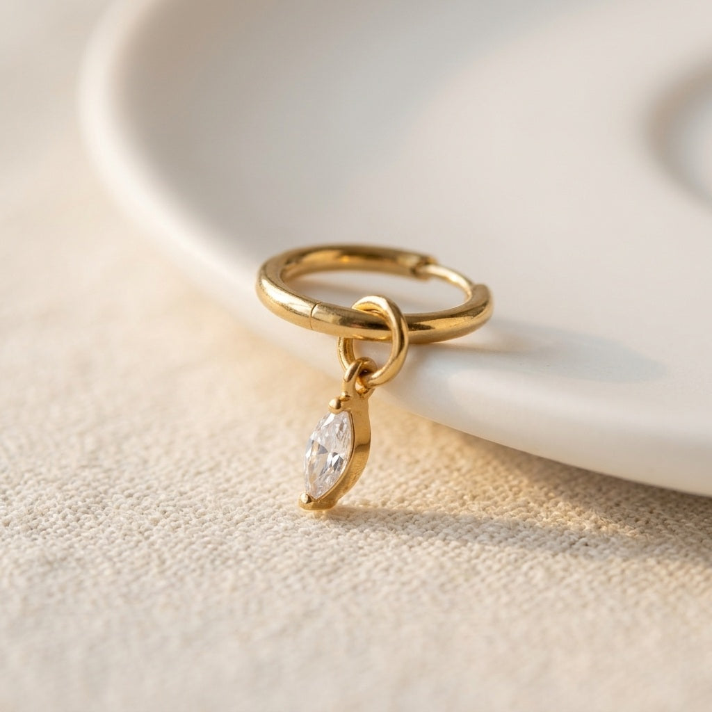 A gold huggie ring with a dangling marquise-cut clear gemstone, resting on a white ceramic dish over a textured beige fabric surface.