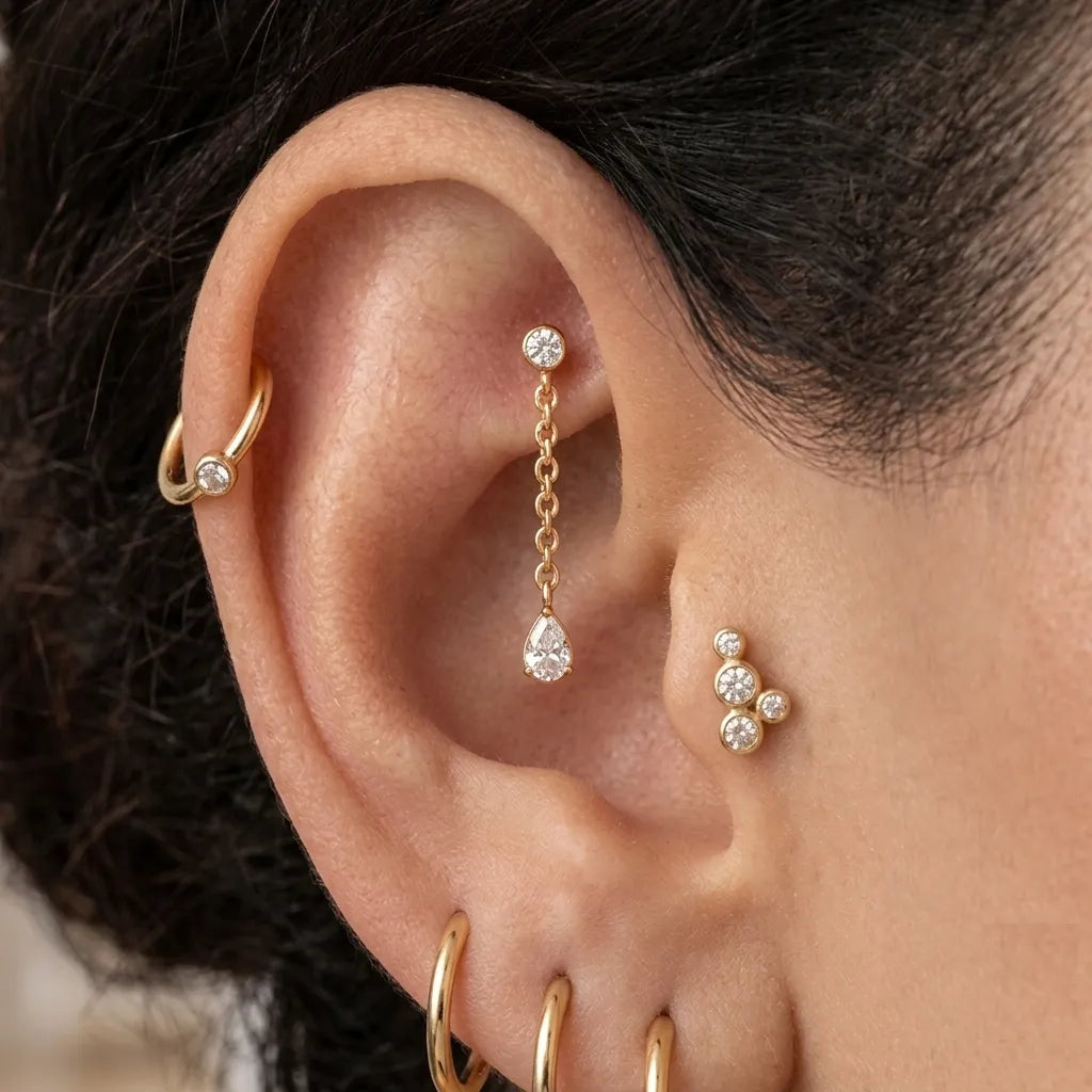 Close-up of a person's ear showcasing multiple gold ear piercings, including a hoop in the cartilage, a dangling chain with a teardrop crystal in the lobe, a small cluster of three crystal studs on the helix, and stacked hoops in the lobe.