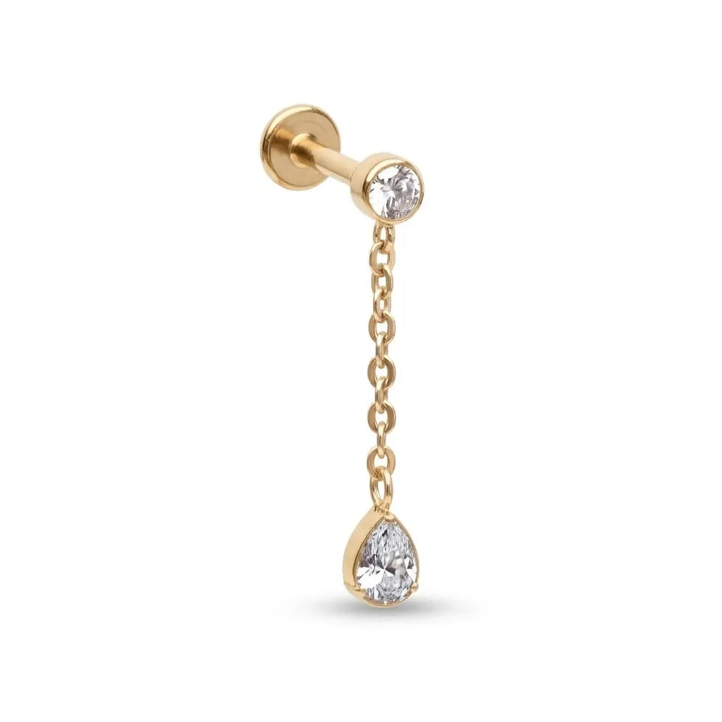 A gold labret stud earring featuring a small clear teardrop gemstone suspended by a delicate chain from a larger round clear gemstone, set against a plain white background.
