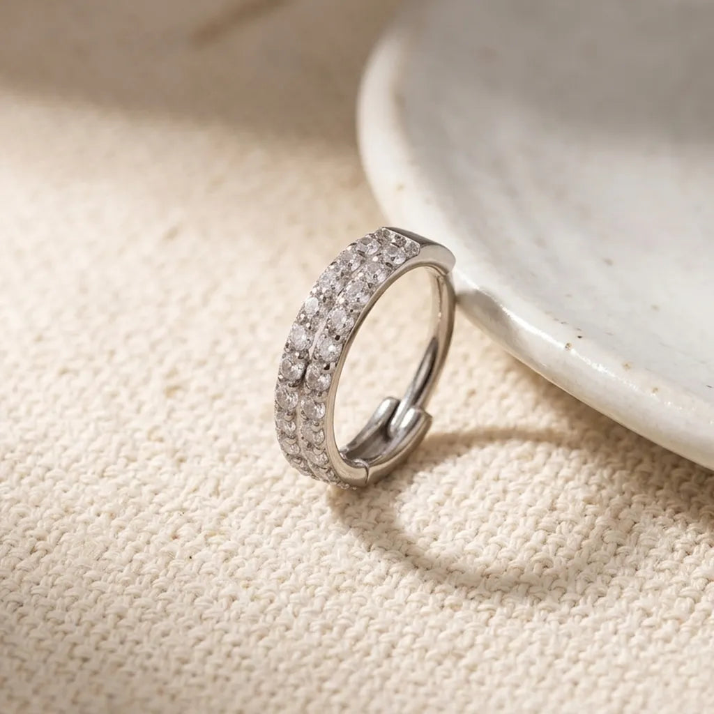 A silver huggie ring with rows of clear cubic zirconia stones, resting on a textured beige fabric next to a white ceramic dish.