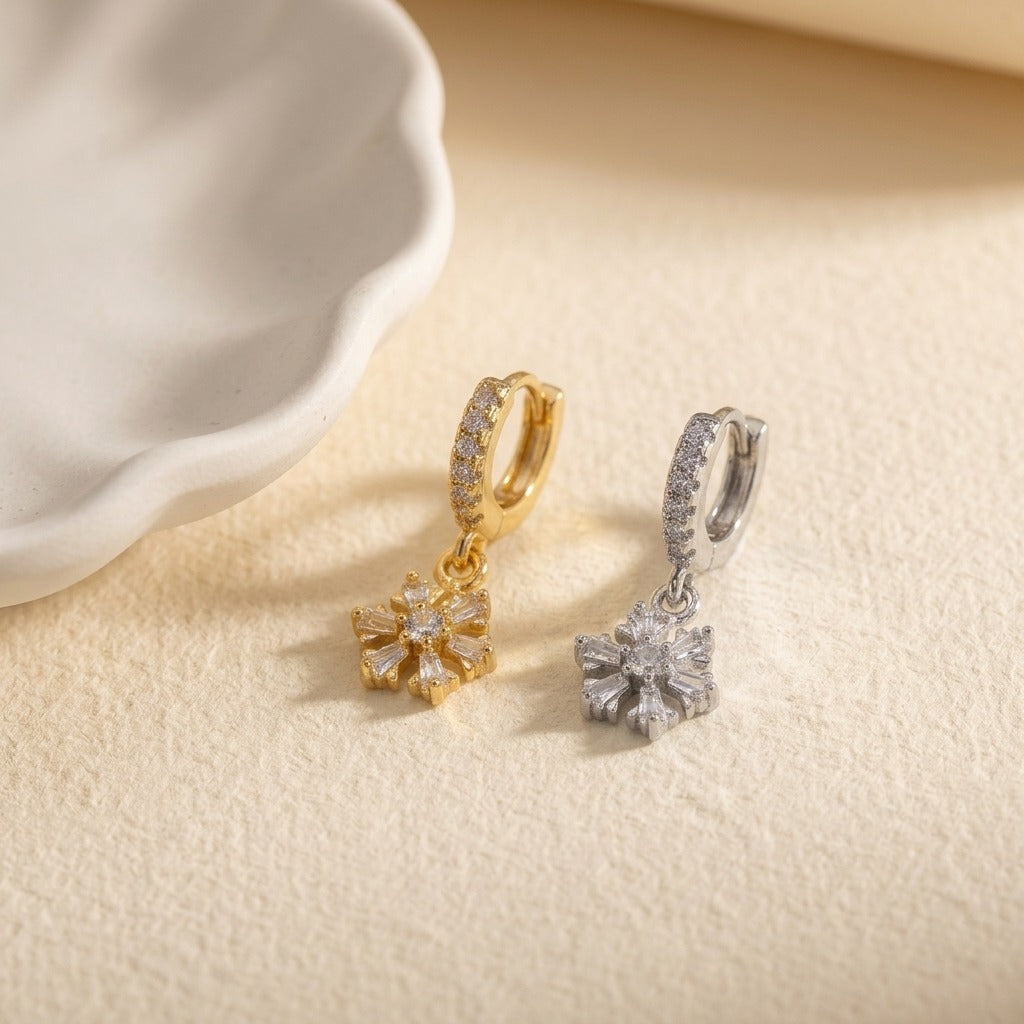 Two small hoop earrings with dangling flower-shaped crystal charms, shown on a warm beige textured surface next to a white dish. One earring is gold-toned and the other is silver-toned.