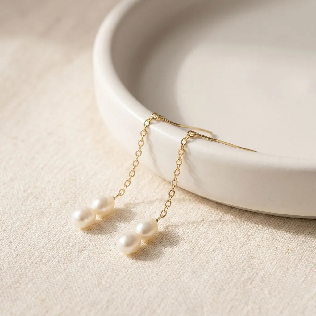 Pearl earrings with gold chains on a beige surface with a white dish in the background