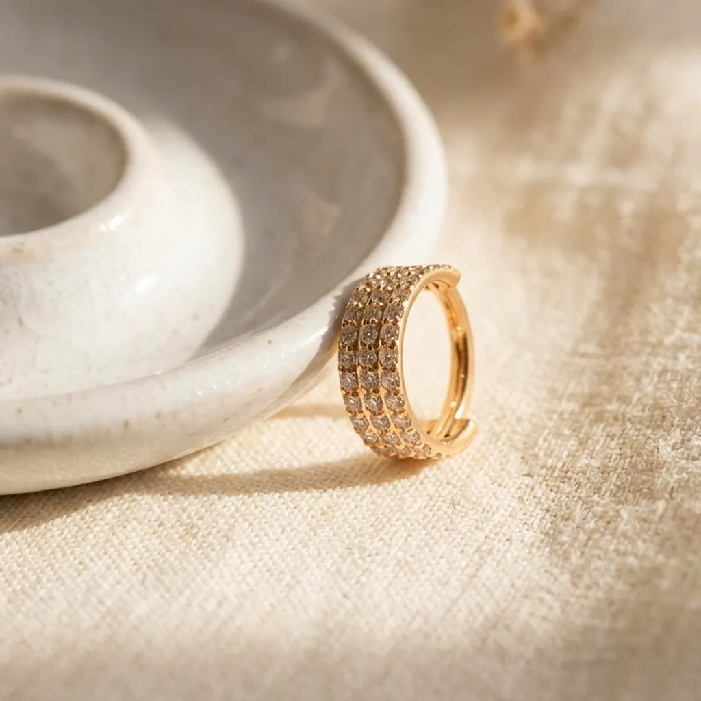 A gold hoop ring with multiple rows of small clear stones, resting on a textured beige fabric next to a white ceramic dish.