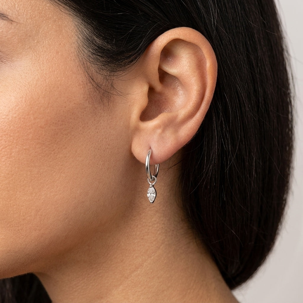 A close-up of a model's ear wearing a small silver hoop earring with a dangling marquise-cut clear gemstone.