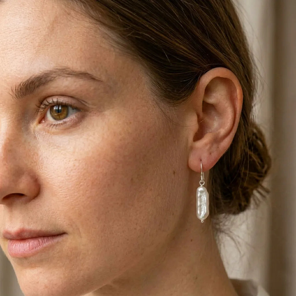 A close-up side profile of a woman wearing a long sterling silver drop earring featuring two round white pearls connected by a delicate chai