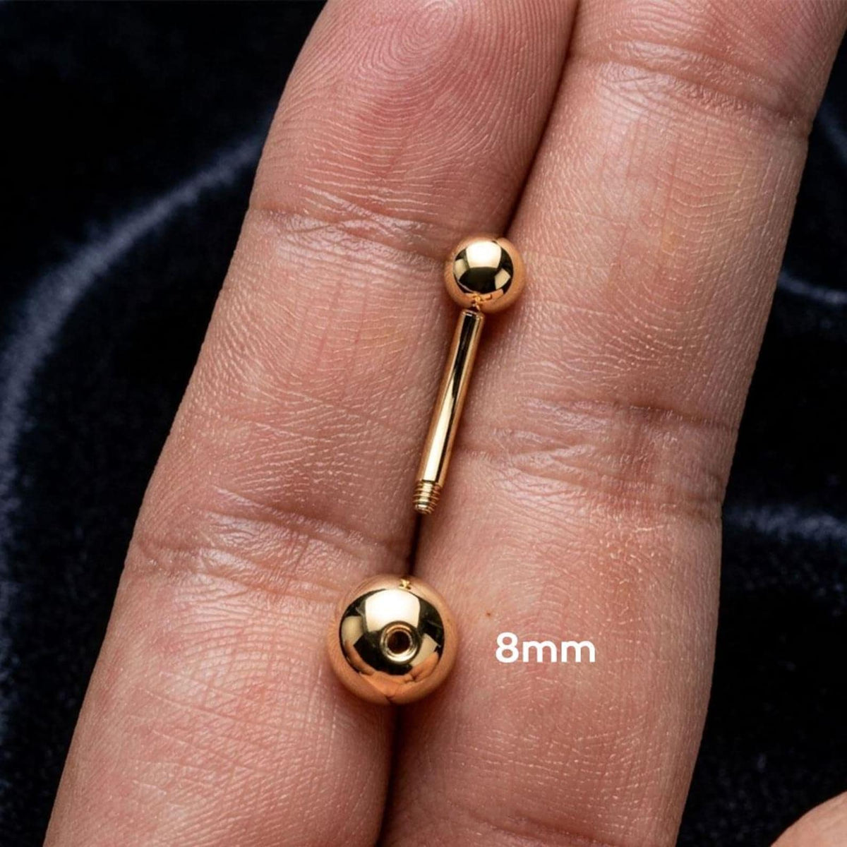 A close-up of a gold barbell nipple piercing jewelry piece resting on a person's finger, with an '8mm' size overlay. The jewelry features a polished ball stud on each end of a straight bar.