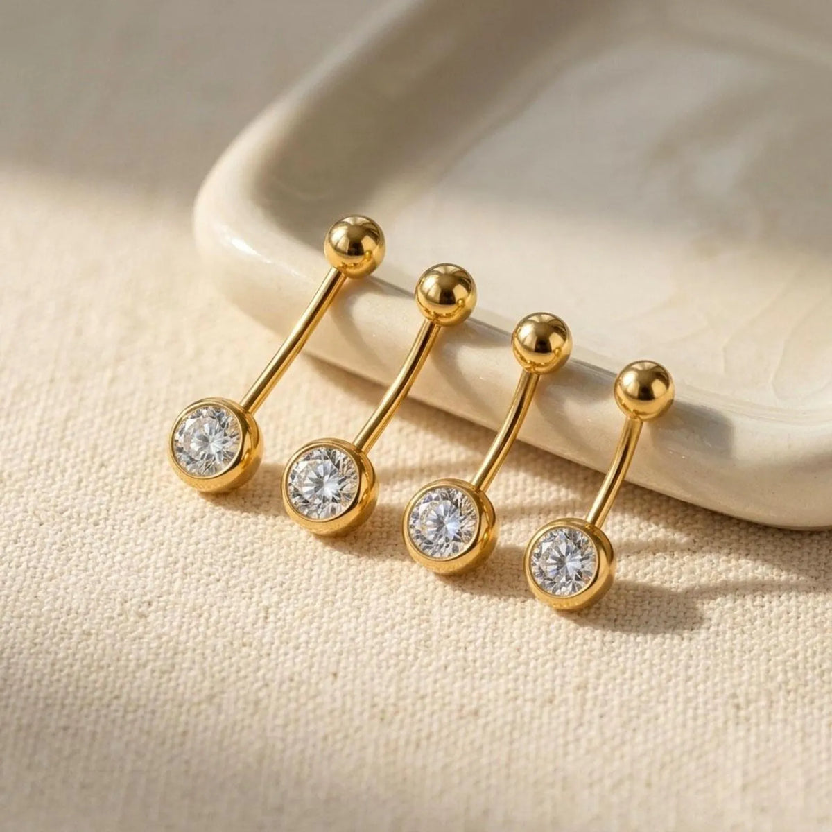 Four gold barbell belly button piercings with round clear gemstones, arranged on a textured beige fabric next to a white ceramic dish.