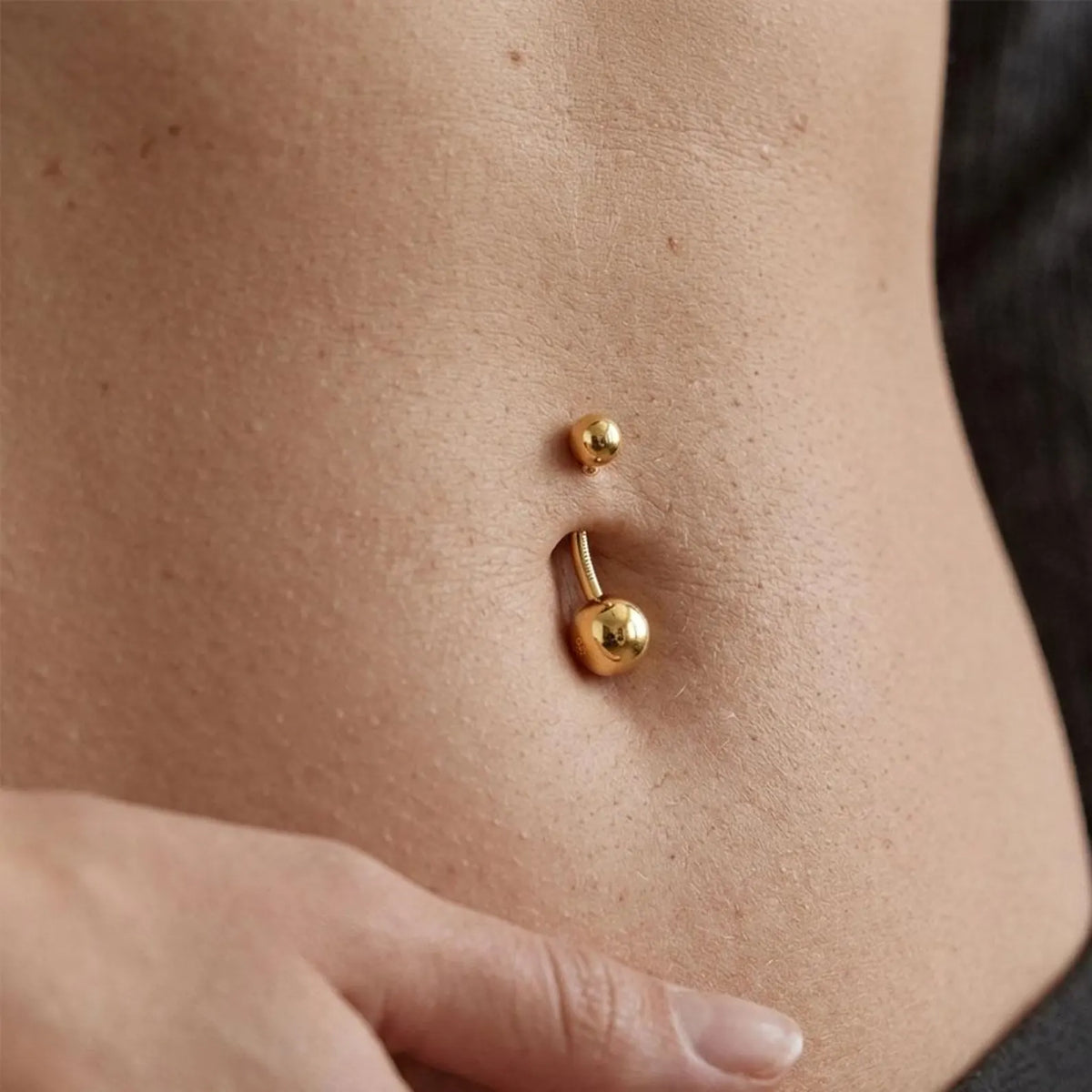 A close-up of a person's lower torso showing two gold belly button piercings: a smaller single bead stud above and a larger curved barbell with a round ball stud below, worn on skin.