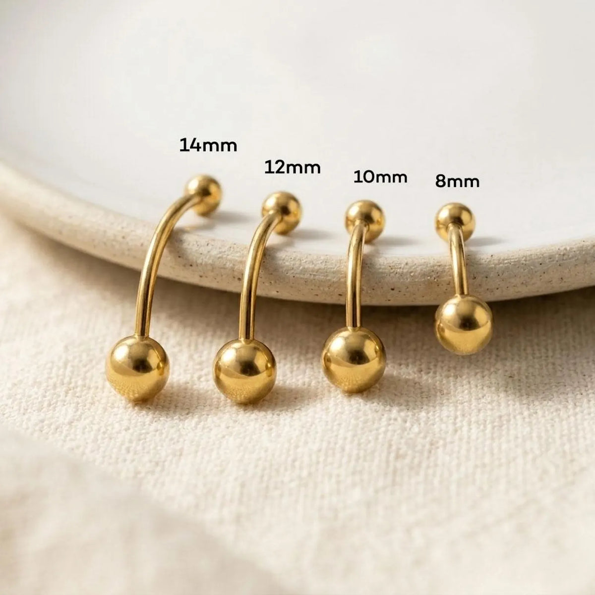 Four gold-colored ball-barbell piercings in different sizes (14mm, 12mm, 10mm, 8mm) arranged on a textured beige fabric surface with a ceramic plate in the background.