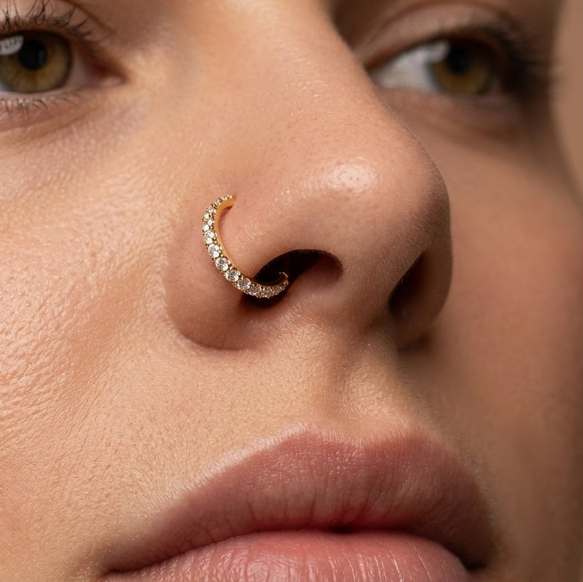 Close-up of a person's nose wearing a small gold crescent-shaped nose stud adorned with clear stones.