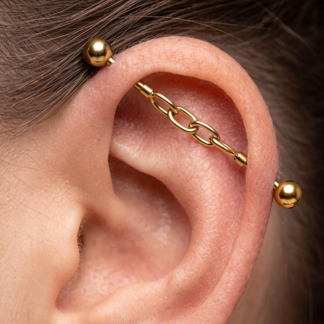 Close-up of a person's ear showcasing a gold ear cuff with a central chain link design, flanked by two gold helix studs.