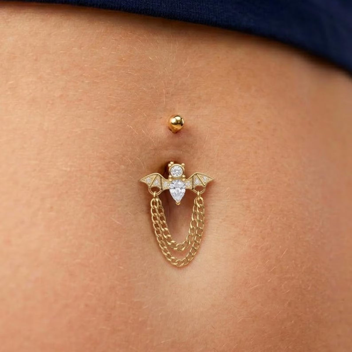 A gold belly button ring with a winged cherub design, a clear heart-shaped crystal, and a hanging chain, shown worn on a person's abdomen.
