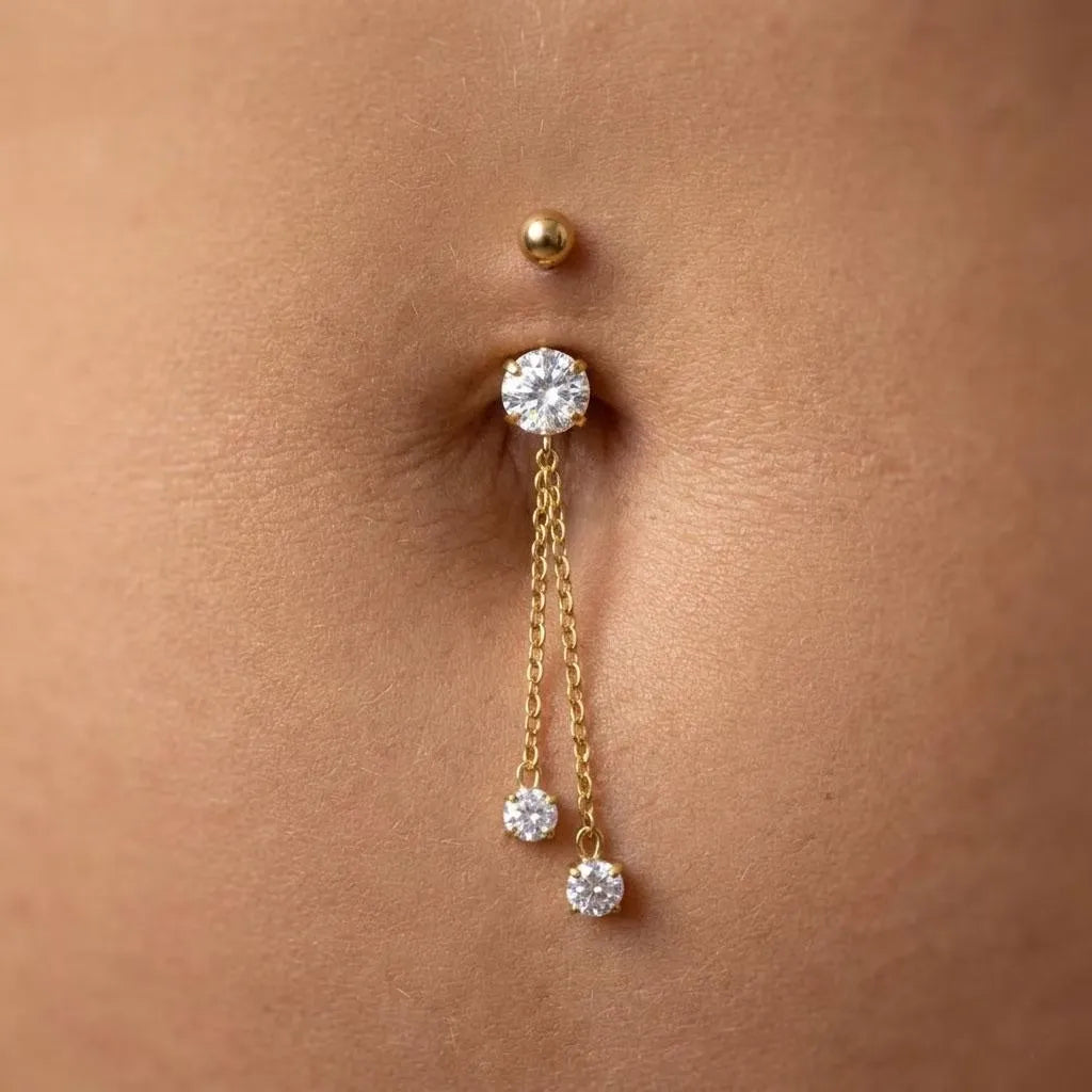 A close-up of a gold belly button ring with a round clear gemstone, a dangling chain with two smaller clear gemstones, and a small gold dot stud above it, shown on skin.