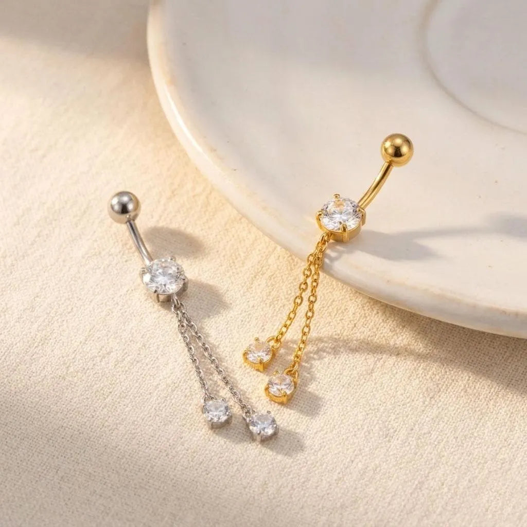 Two dangling belly button rings displayed on a light beige textured surface with a white ceramic plate in the background. One ring is silver-toned with a single clear stone and three dangling chain charms with clear stones.