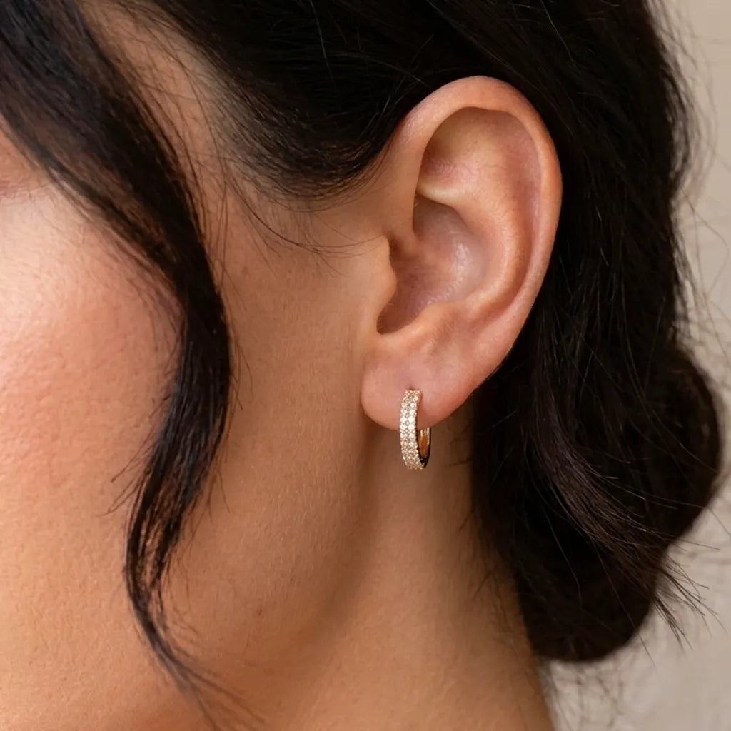 A close-up of a person's ear wearing a small gold huggie hoop earring set with clear pavé stones, against a soft light beige background.