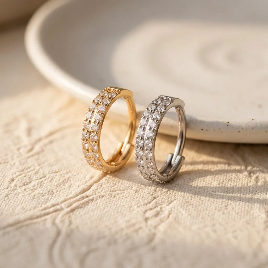 Two small hoop earrings with rows of clear stones, one in yellow gold and one in silver, resting on a textured beige fabric surface with a ceramic dish in the background.