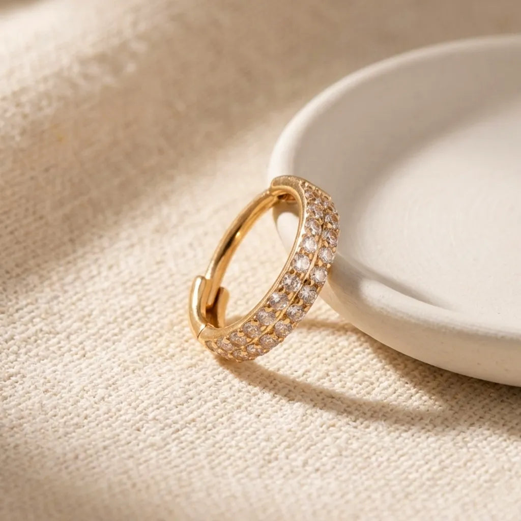 A gold huggie hoop ring adorned with rows of small clear pavé stones, resting on a textured beige fabric next to a white ceramic dish.