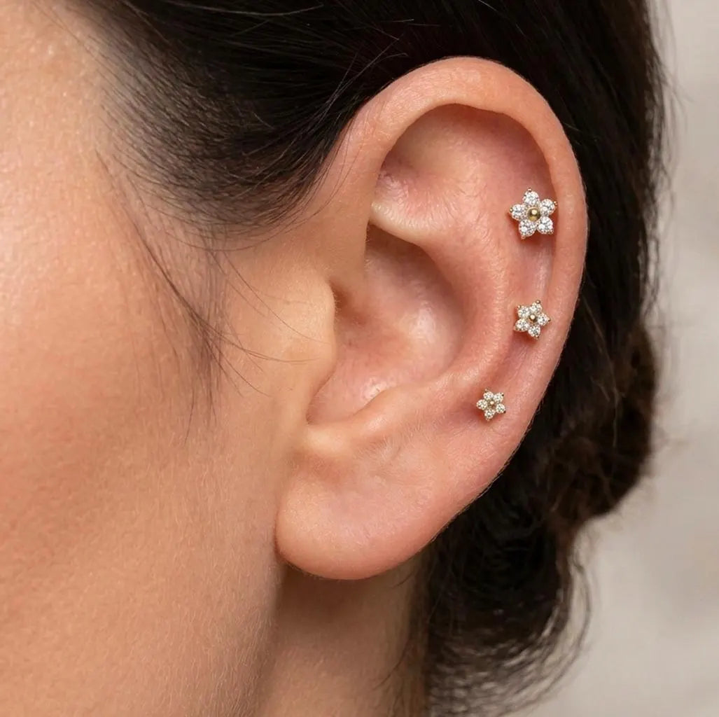 Three small flower-shaped stud earrings with clear stones worn on a person's ear, shown in a close-up lifestyle shot.