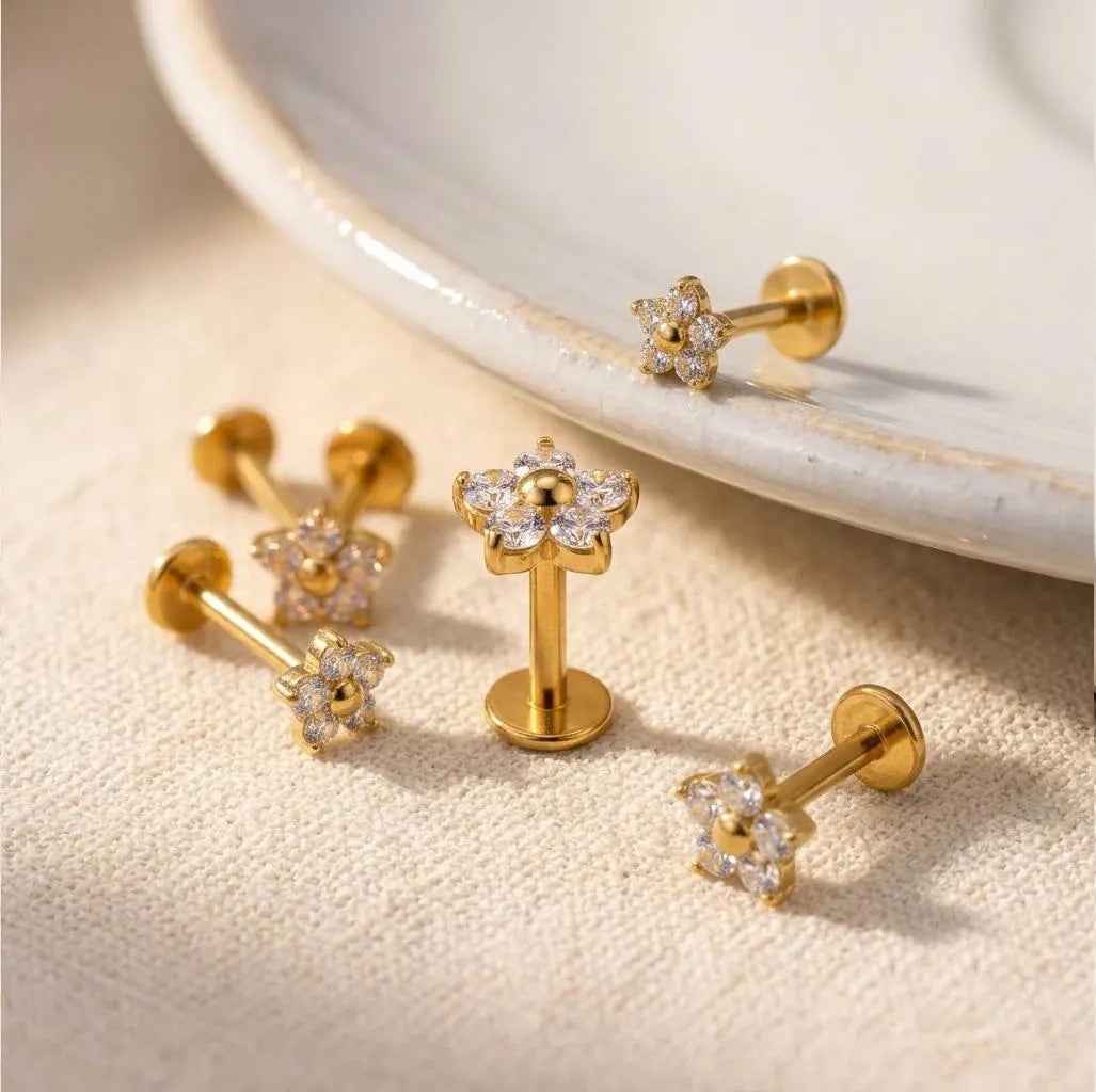 Five gold floral stud earrings with clear crystal-like accents are arranged on a textured beige fabric surface, with one earring resting on the edge of a white ceramic dish in the background.