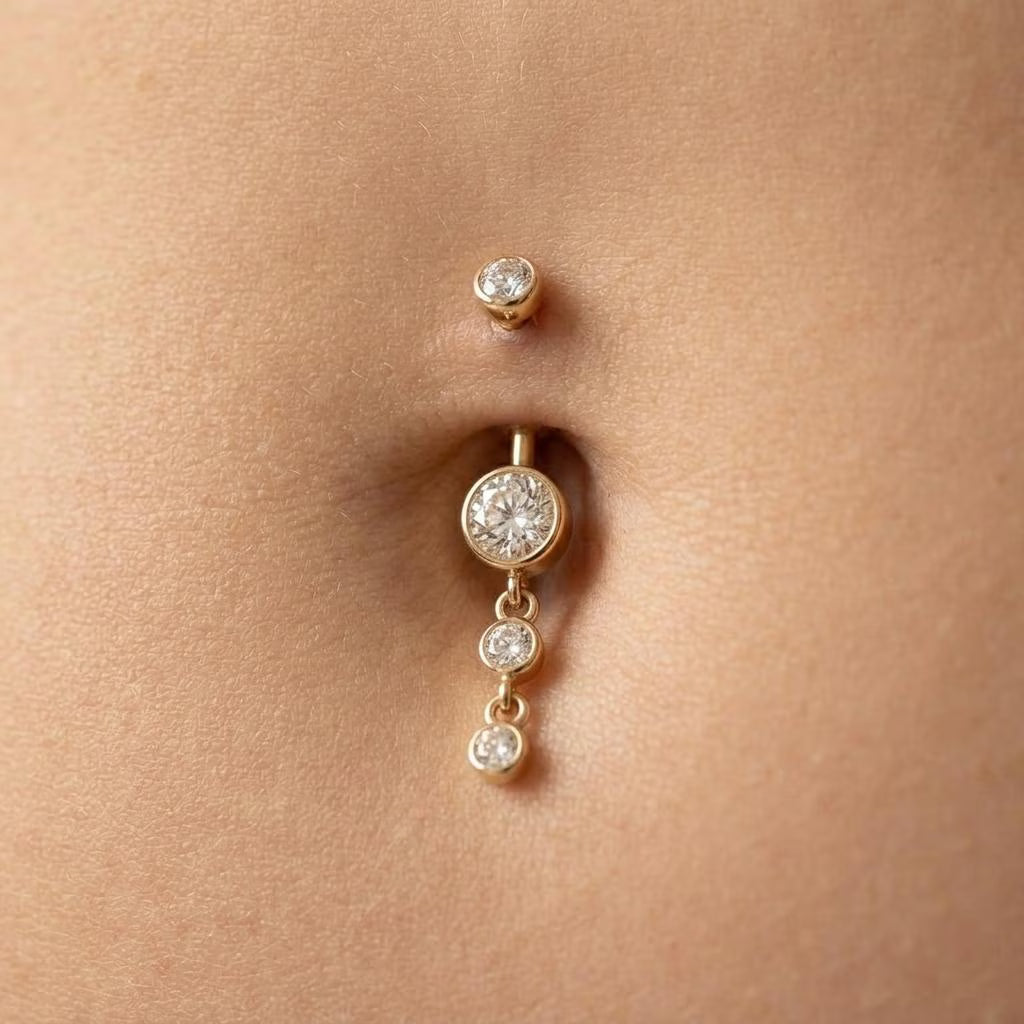 A gold belly button ring with a dangling barbell featuring three clear round gemstones, shown worn on a person's abdomen.