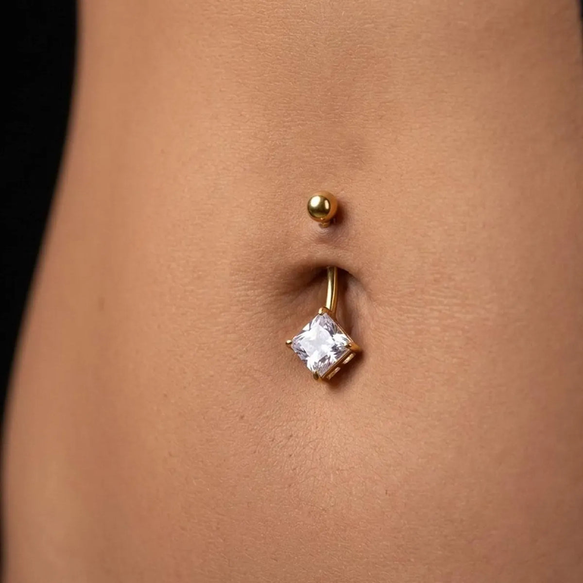 A close-up of a person's lower abdomen showing a gold navel piercing with a square clear gemstone dangling below a small gold bead.