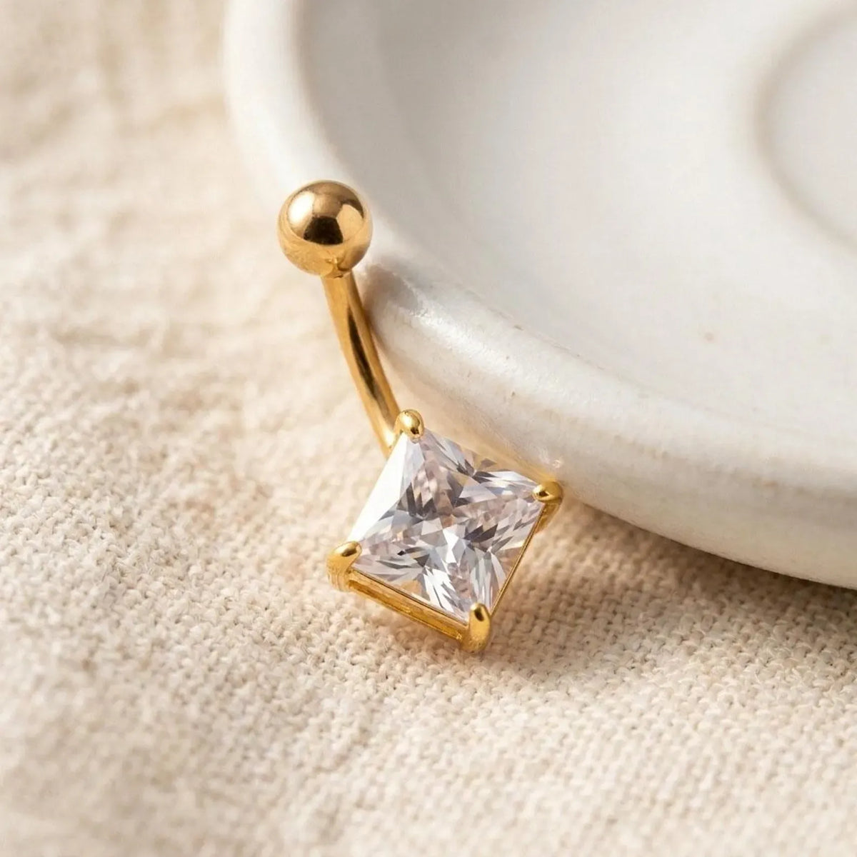 A gold-plated curved barbell belly button ring with a clear princess-cut cubic zirconia gemstone, resting on a textured beige fabric next to a white ceramic dish.