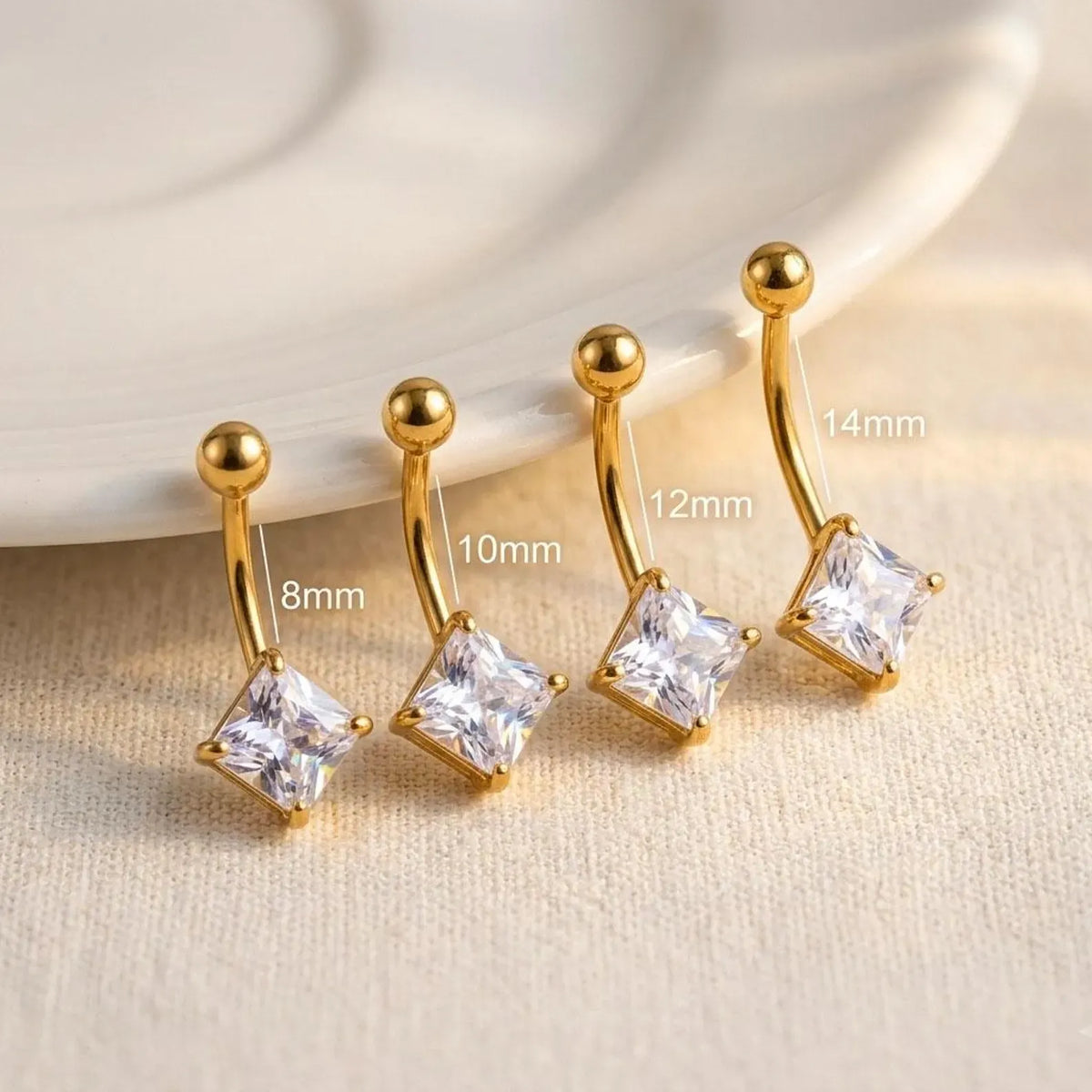 Four gold-plated belly button rings with clear square gemstones, shown in sizes 8mm, 10mm, 12mm, and 14mm, with size labels overlaid on a beige fabric background next to a white ceramic plate.