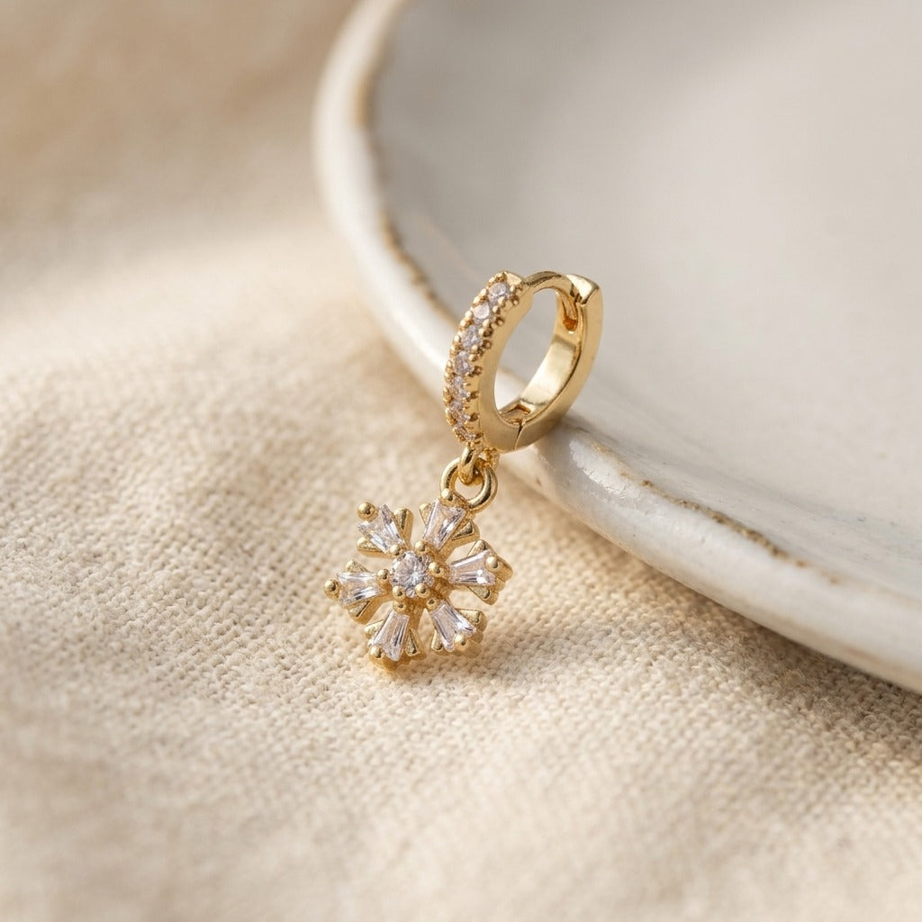 A gold huggie hoop earring with a dangling flower-shaped crystal charm, displayed on a textured beige fabric next to a white ceramic dish.