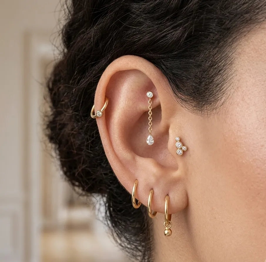 A close-up of a person's ear showcasing multiple gold and silver cartilage piercings, including hoop earrings, a dangling chain, and stud earrings, with dark hair framing the ear.