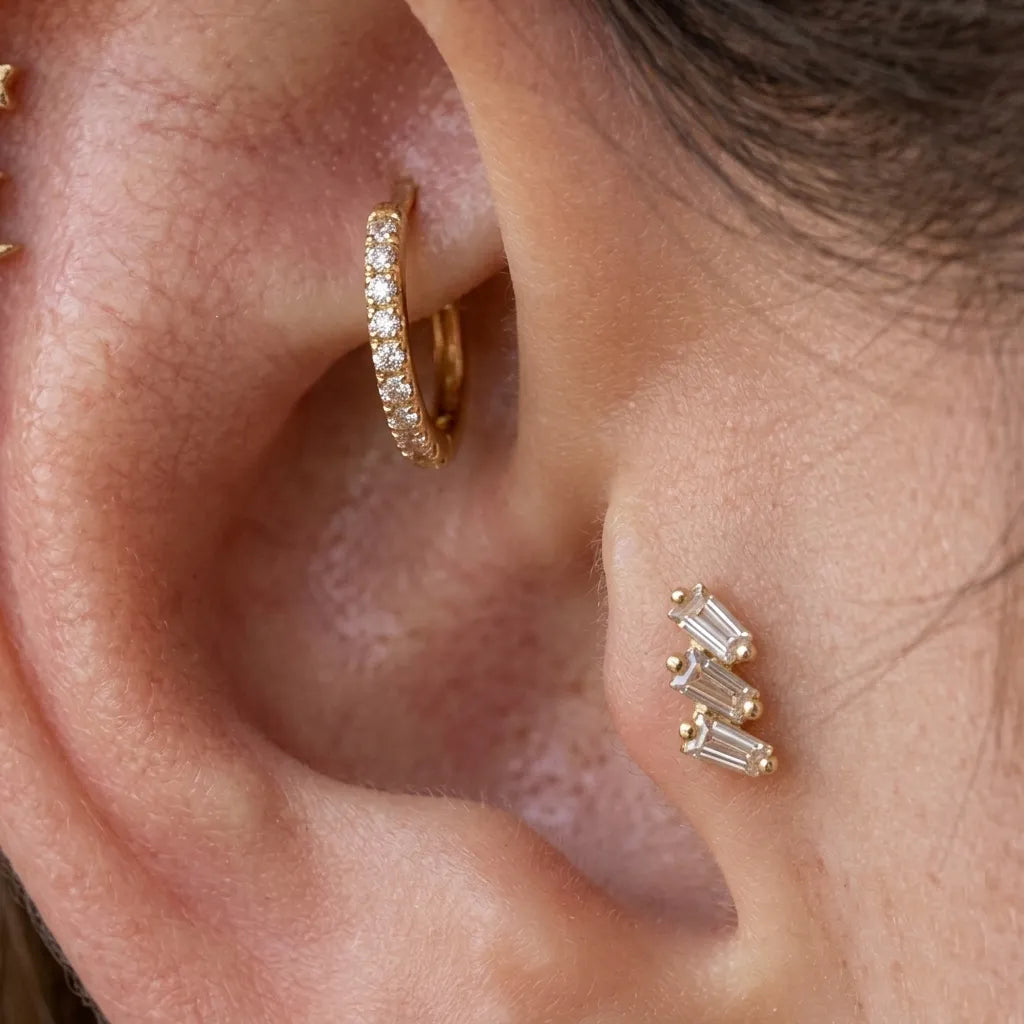 Close-up of an ear showing two gold ear piercings: a small huggie hoop with clear stones on the upper ear cartilage and a cluster of three gold baguette stud earrings on the lobe.