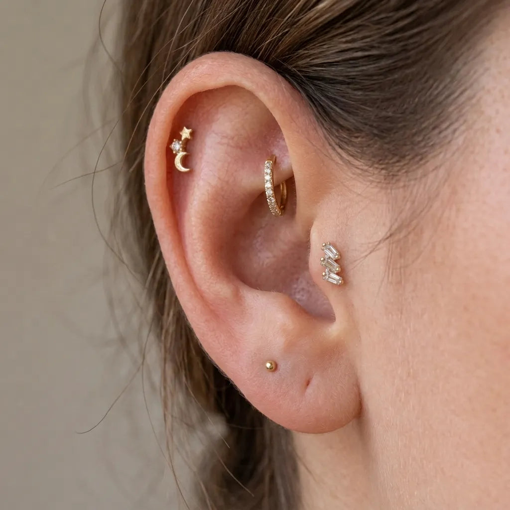 Close-up of a person's ear showcasing multiple gold and silver ear piercings, including a triple cartilage stack with stars and a crescent moon, a huggie hoop, a small cluster, and a single pearl stud.