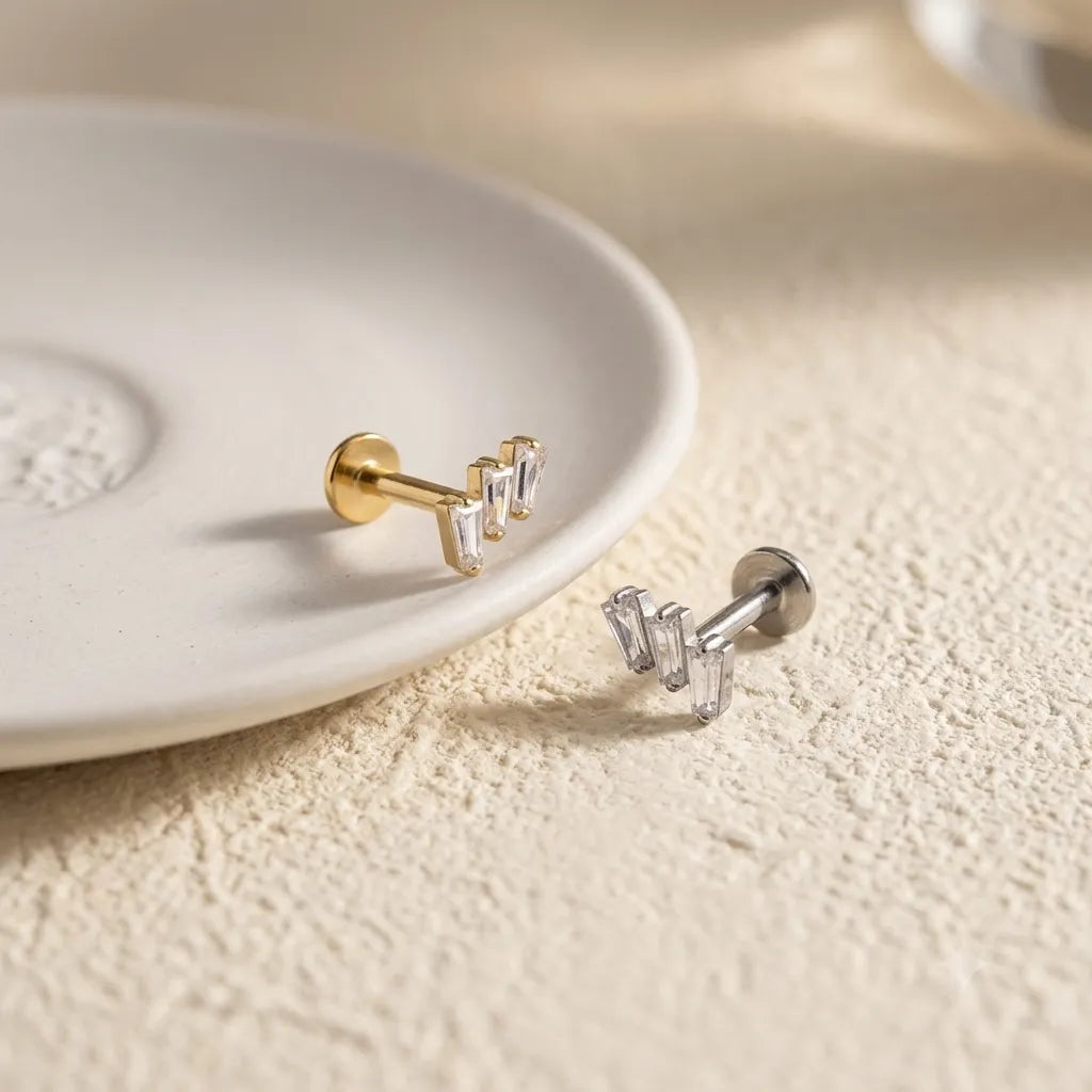 Two small geometric stud earrings resting on a white ceramic dish, one in polished gold and one in polished silver, on a textured beige surface.