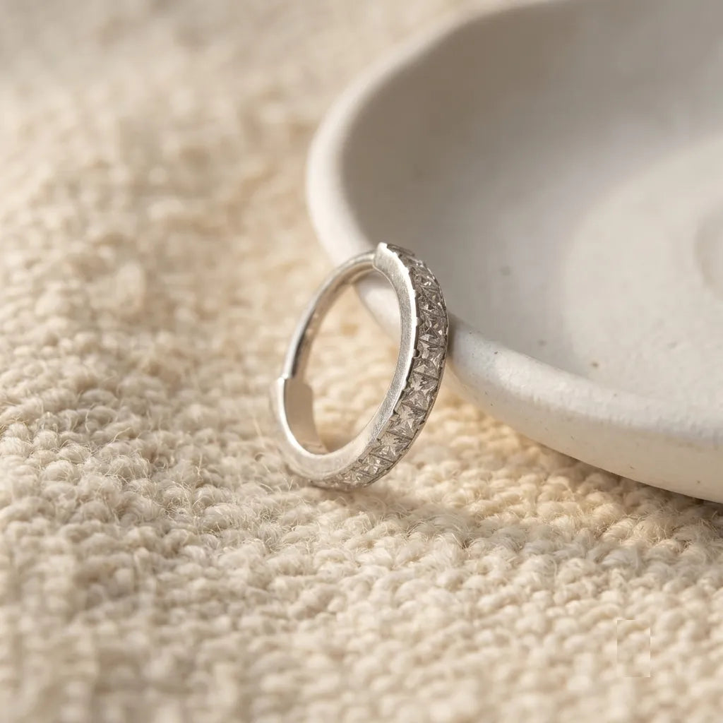 A silver hoop ring with a row of small clear stones, resting on a textured beige fabric next to a light ceramic dish.