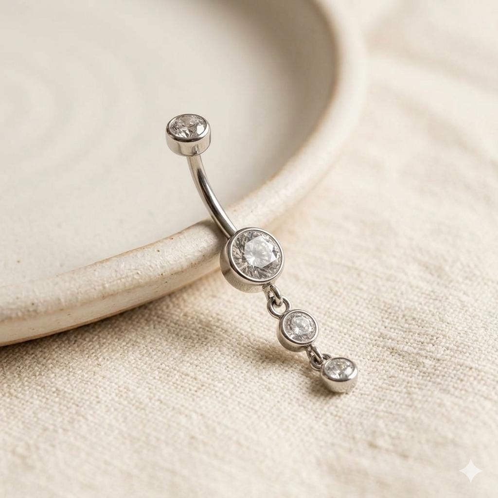 A silver-toned curved barbell belly button ring with three dangling clear gemstones, photographed on a textured beige fabric next to a ceramic dish.