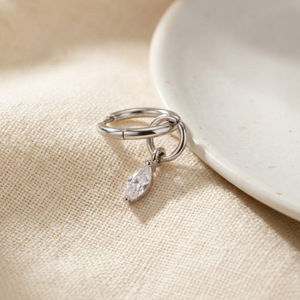 A silver huggie hoop earring with a dangling marquise-cut clear gemstone, resting on a textured beige fabric next to a white ceramic dish.