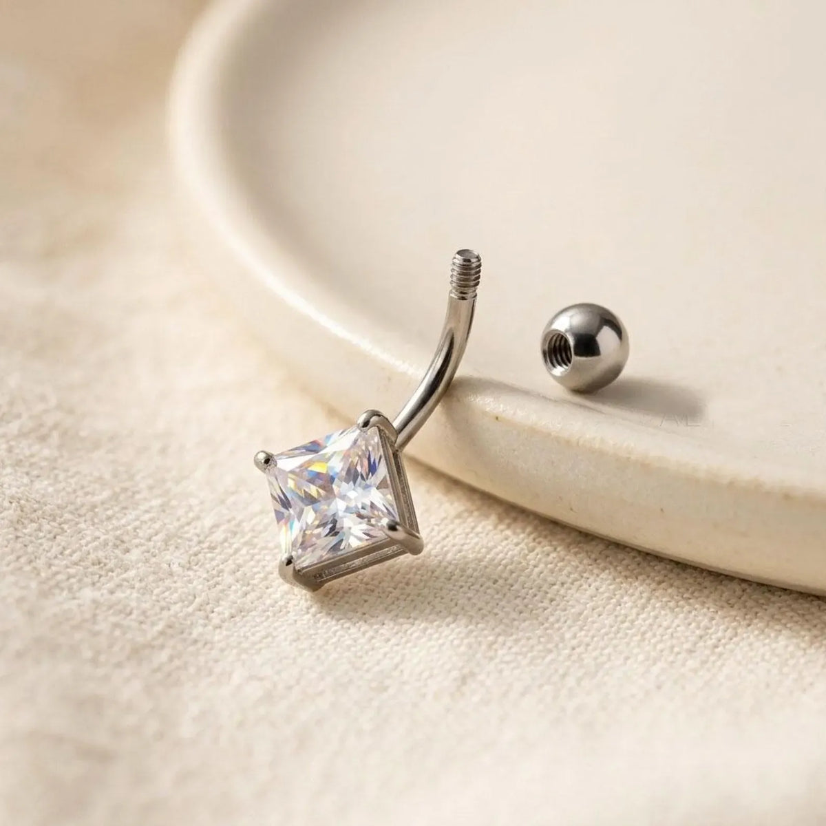 A silver-toned curved barbell piercing stud with a clear princess-cut gemstone, shown next to its matching ball-back screw on a textured beige fabric surface.