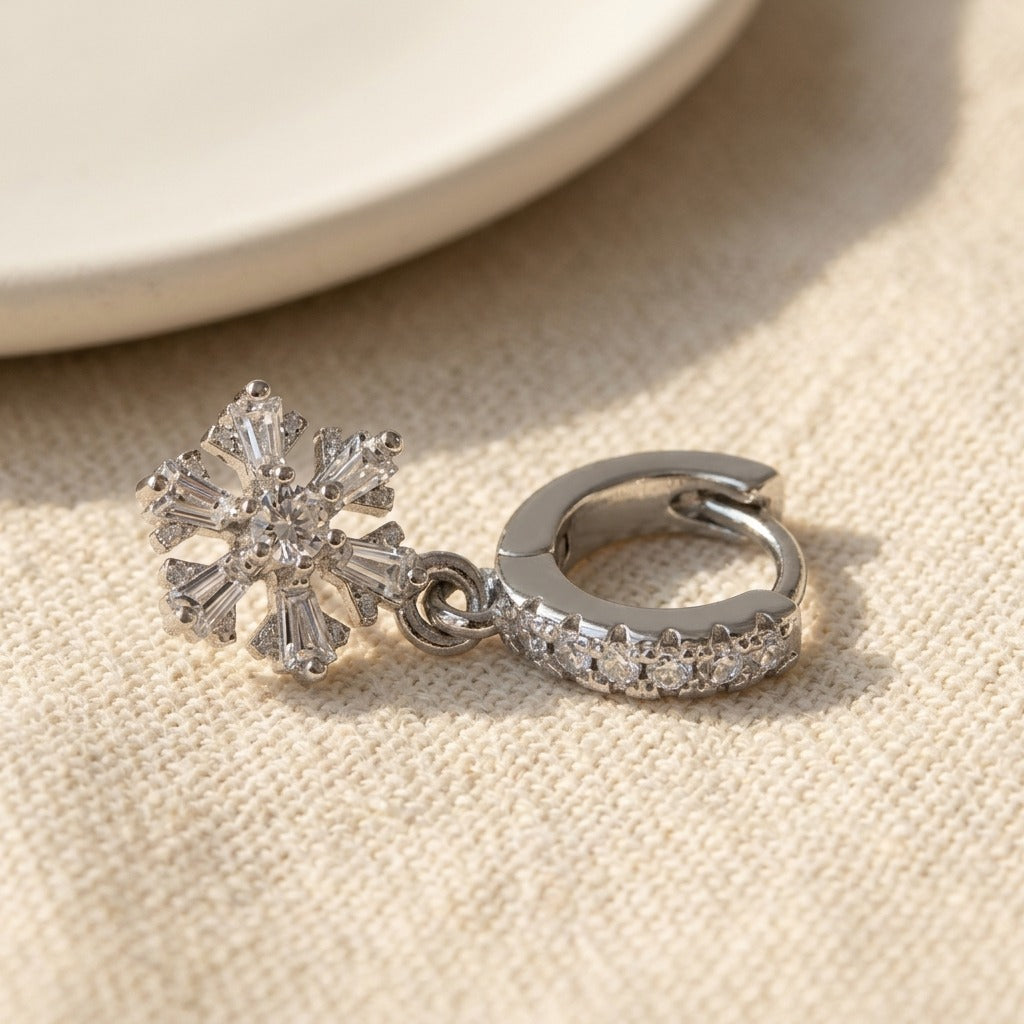 A silver-toned huggie hoop earring with a dangling snowflake charm, both adorned with clear crystals, resting on a textured beige fabric surface.