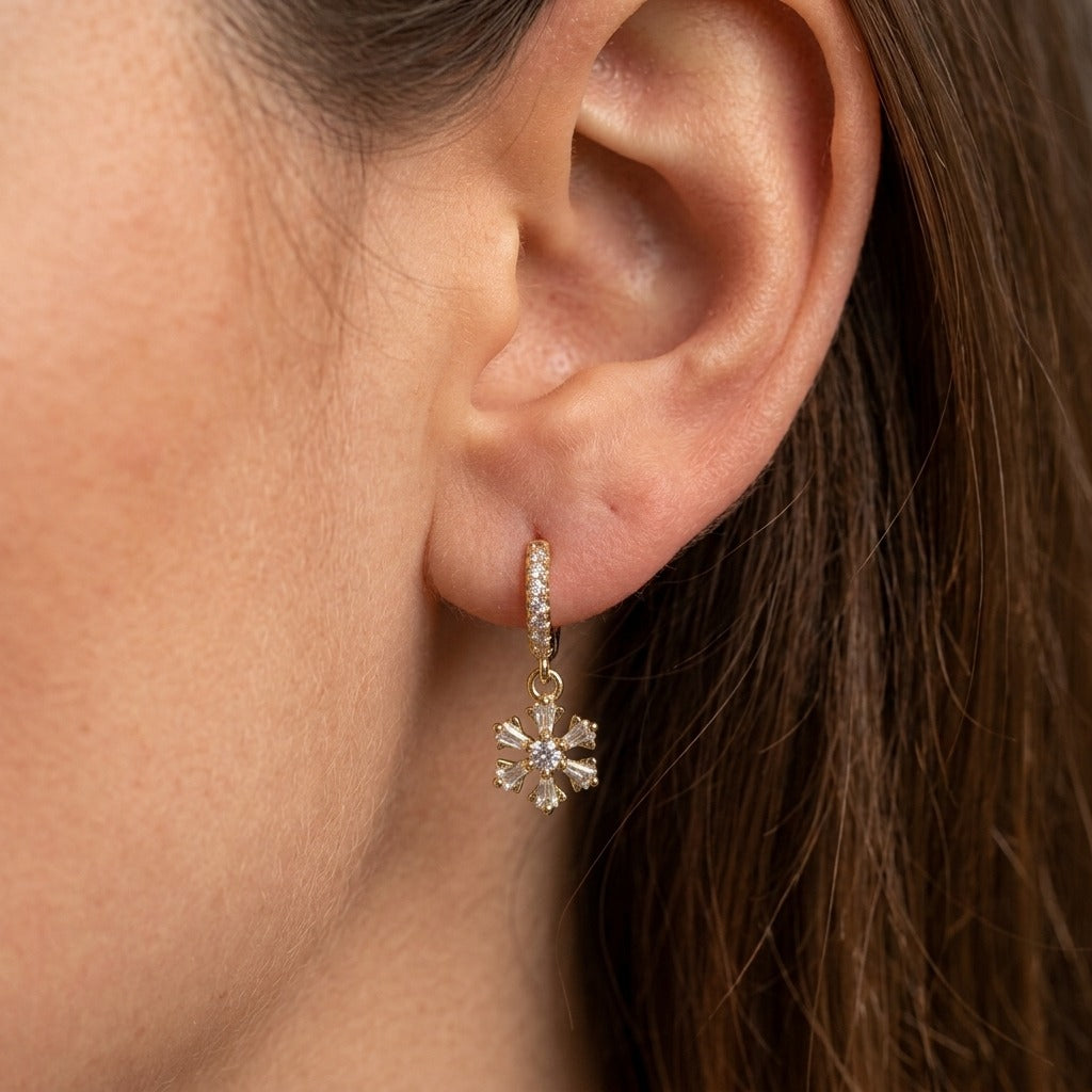 A close-up of a model's ear wearing a small gold huggie hoop earring with a dangling flower charm set with clear stones.