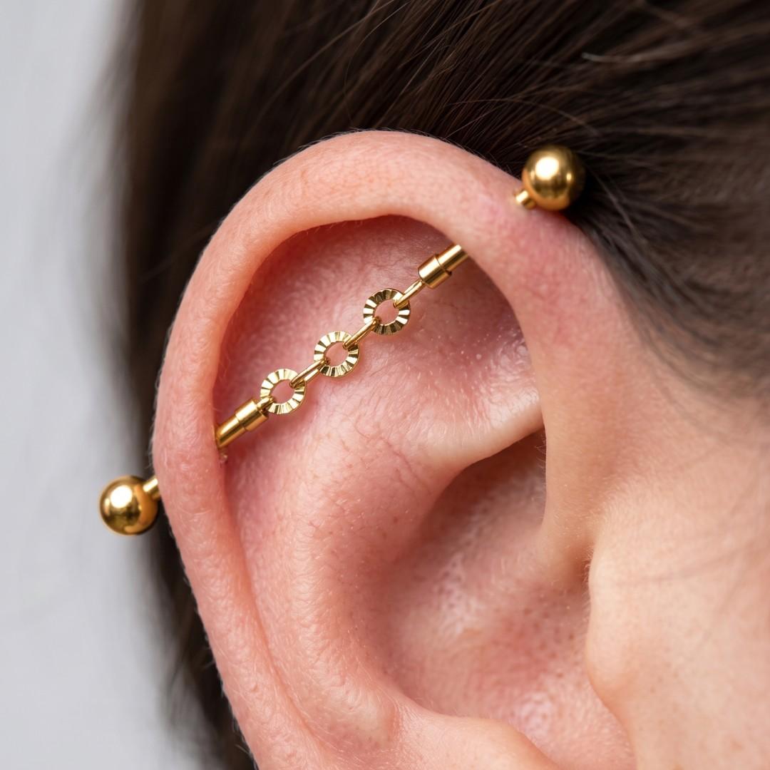 A close-up of a person's ear showcasing a gold-colored multi-ring cartilage barbell ear cuff with three decorative circular links and two ball studs.