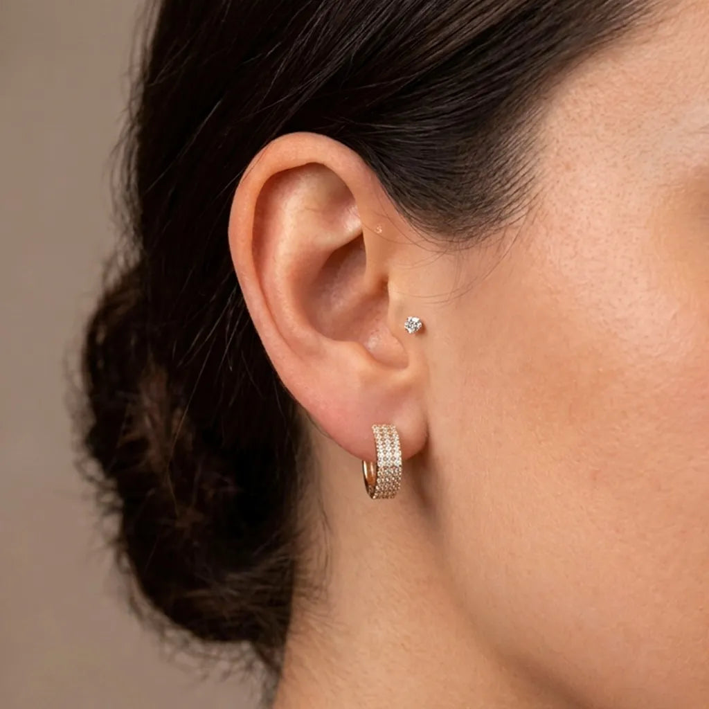 Close-up of a woman's ear wearing a small gold hoop earring set with clear stones and a tiny matching stud earring, against a soft beige background.
